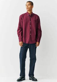 Bordeauxrode overhemd met lange mouwen, gecombineerd met donkere spijkerbroek. Het shirt heeft een klassieke kraag en witte knopen.