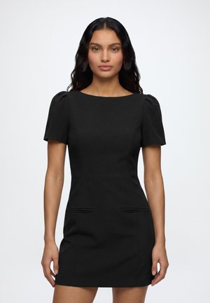 Frau mit langen dunklen Haaren, die ein eng anliegendes schwarzes Kurzarmkleid mit rundem Ausschnitt trägt, steht vor einem einfarbigen hellen Hintergrund.