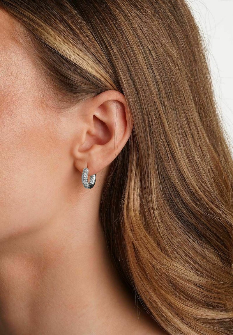 Kvinnas öra med medellång ljusbrun hår som bär en liten silver hoop-örhänge prydd med klara kristaller.