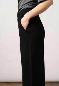 Zwarte, wijde broek van gladde stof, met een zijzak en een strakke, gestroomlijnde silhouet. Van opzij getoond.