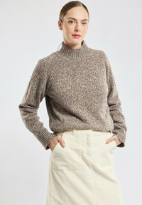 Femme avec les mains dans les poches portant un pull en laine marron et une jupe en velours côtelé crème, se tenant contre un fond uni.