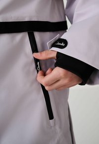 Svetlo šedá bunda s čiernymi akcentmi, vybavená zipsom v oblasti pásu a logom na manžete rukáva. Hladký, vodoodolný materiál.