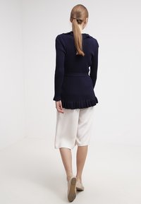 Námořnicky modrý žebrovaný svetr s kapucí a volánkovým lemem. Kombinován s krémovými, volně střiženými kalhotami culottes a béžovými botami.