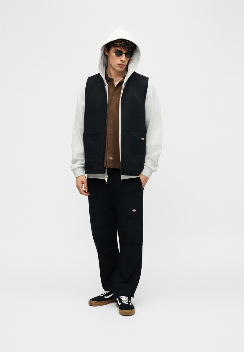 Giovane uomo con indosso una felpa grigia con cappuccio sotto un gilet nero senza maniche, camicia marrone con bottoni, pantaloni cargo neri, scarpe da ginnastica nere e occhiali da sole rotondi.