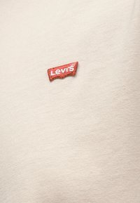 Μικρό κόκκινο λογότυπο Levi's ραμμένο σε κρεμ ύφασμα, που δείχνει λεπτομερή υφή του υλικού και της ραφής.