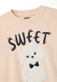 Roze sweatshirt met zwarte tekst die "SWEET" zegt. Bevat een pluche witte hond applicatie met een zwarte vlinderdas en roze ooraccenten.