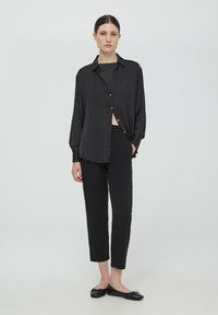 Camicia nera in satin con bottoni, indossata con pantaloni neri aderenti e ballerine nere. La camicia ha una vestibilità ampia e un orlo arrotondato.