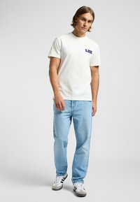 Hvid bomulds t-shirt med et blåt "LEE" logo på brystet, parret med lyseblå denim jeans og hvide sneakers. Enkel, afslappet design.