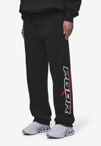 Svarta sweatpants med en avslappnad passform, med vita och rosa logotyper längs sidan och en mjuk yta. Parat med ljusgrå sneakers.