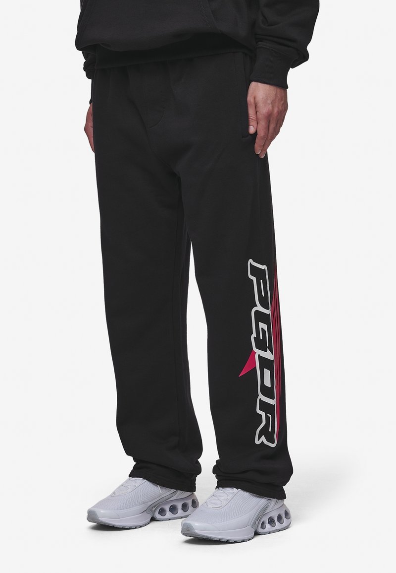 Svarta sweatpants med en avslappnad passform, med vita och rosa logotyper längs sidan och en mjuk yta. Parat med ljusgrå sneakers.