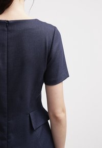 Mörkblå strukturerad klänning med korta ärmar, ett subtilt mönster och en dragkedja bak med en liten slits i midjan.