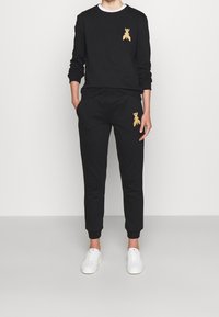 Felpa nera con pantaloni coordinati, entrambi con un accento ricamato in oro; presenta un collo rotondo e polsini a costine sui polsi e sulle caviglie.