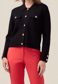 Svart stickad cardigan med krage, två framfickor och guldknappdetaljer. I kombination med figurnära korallfärgade byxor. Enfärgad bakgrund.