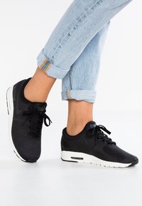 Mustat Nike-tossut, joissa on teksturoitu päällinen, valkoinen pohja ja perinteinen nauhoitus. Farkut käännetty lahkeista, näyttäen nilkan.