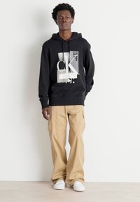 Felpa nera con stampa grafica, cappuccio con lacci e polsini a coste. Abbinata a pantaloni cargo khaki e sneaker bianche, in piedi contro uno sfondo semplice.