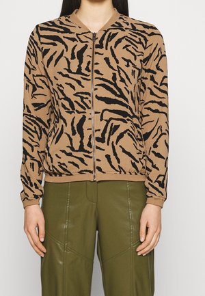 Femme portant une veste zippée marron et noire à rayures tigre, associée à un pantalon taille haute vert olive.
