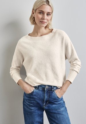Maglione in maglia beige chiaro con scollo ampio e polsini a costine, abbinato a jeans in denim blu con vestibilità classica e tasche.