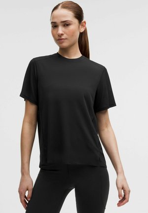 Femme aux cheveux attachés, portant un t-shirt noir uni à manches courtes et des leggings noirs, debout devant un fond clair uni.