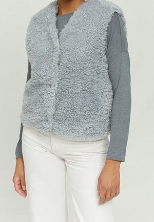 Personne portant une veste sans manches duveteuse gris clair sur une chemise grise à manches longues et un pantalon blanc, debout devant un fond uni.
