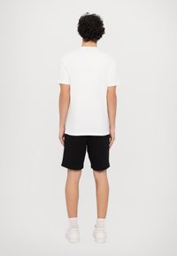 Hvid kortærmet t-shirt, afslappet pasform, parret med sorte shorts. Modellen står med ryggen til kameraet, iført hvide sneakers og lyse sokker.