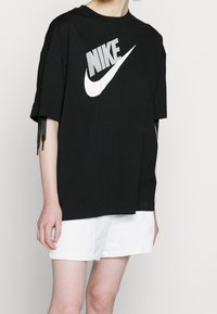 Henkilö, jolla on päällään löysä musta Nike T-paita valkoisella logolla ja valkoiset tekstuurilla varustetut shortsit, seisoo yksinkertaista vaaleaa taustaa vasten.