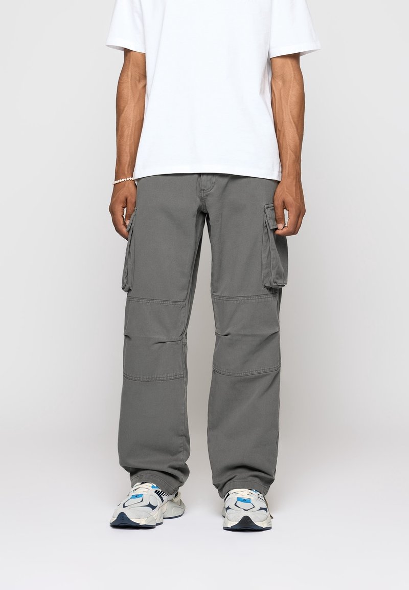 Homme portant un pantalon cargo gris ample, des baskets blanches avec des accents bleus, et un T-shirt blanc uni, se tenant devant un fond clair.