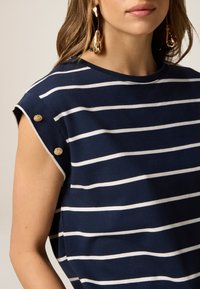 Top a rayas azul marino con rayas blancas. Presenta mangas cortas, acentos de botones dorados en los hombros y una tela suave y ligera.