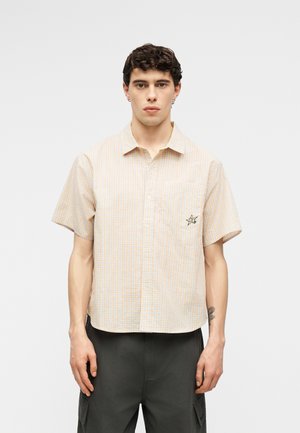 Junger Mann mit kurzen lockigen Haaren, der ein kurzärmeliges beiges kariertes Hemd mit einem Stern-Logo auf der Brusttasche trägt.