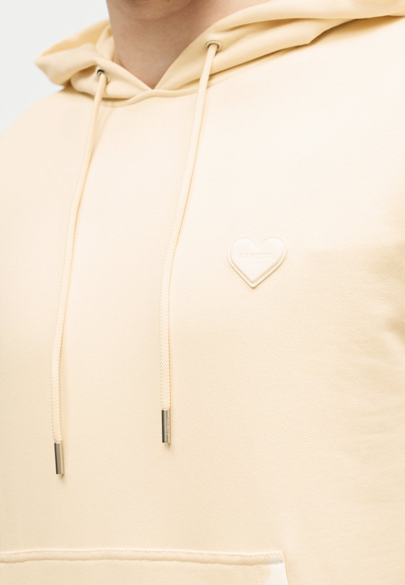 Beige hoodie with drawstrings and a small heart-shaped patch on the chest near the left shoulder.