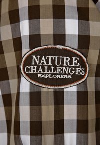 Brązowo-biała tkanina w kratkę z haftowaną owalną naszywką z napisem "NATURE CHALLENGES EXPLORERS", z kontrastującą bordiurą.
