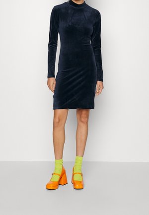 Mujer que lleva un vestido ajustado de terciopelo negro de manga larga con calcetines verde neón y zapatos de tacón alto gruesos de color naranja brillante.