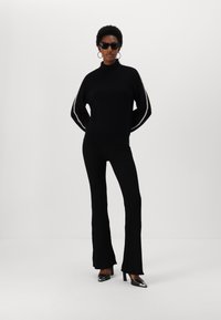 Pulover negru din tricot cu guler înalt, textură ribbată și detalii laterale albe, completat cu pantaloni evazați negri din tricot ribbat și pantofi cu vârf ascuțit.
