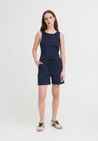 Woman wearing navy sleeveless top and matching drawstring shorts with beige sneakers, standing against a plain white background.