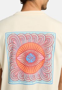 Camiseta beige con un diseño gráfico colorido en la parte trasera. El diseño incluye una forma de ojo con líneas en espiral en rosa, naranja y azul.
