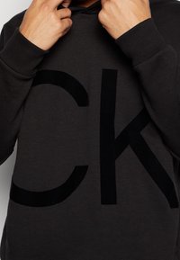 Sudadera con capucha negra con un destacado logotipo grande "CK" en negro sobre un fondo gris oscuro. Fabricada con tela suave, con puños acanalados y capucha con cordón ajustable.