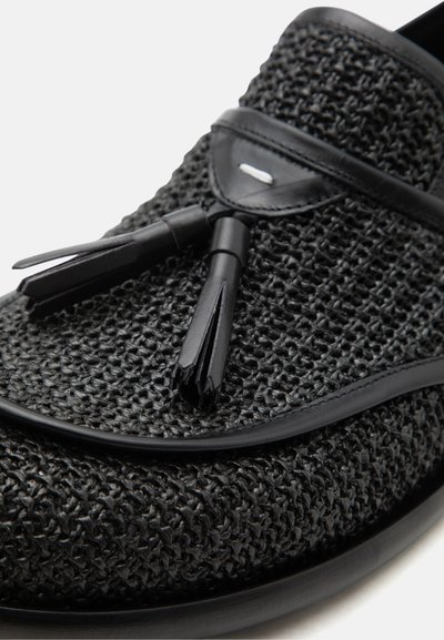 Μαύρο υφαντό δερμάτινο μοκασίνι με μαύρες δερμάτινες φούντες και διακόσμηση σε λευκό φόντο.