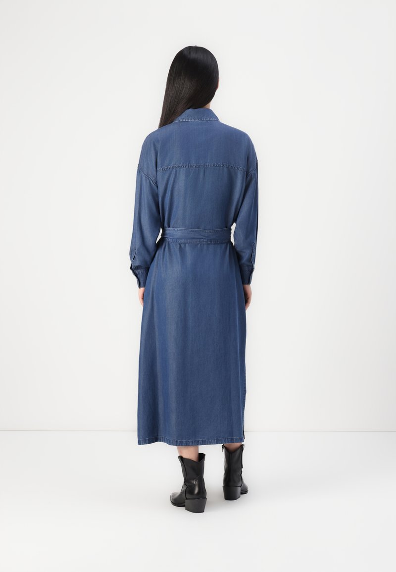 Femme aux longs cheveux noirs, portant une robe midi en denim bleu ceinturée et des bottines noires, se tenant devant un fond blanc uni.