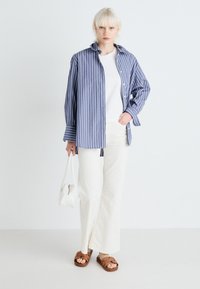 Camicia button-up a righe blu e bianche, indossata sopra una t-shirt bianca. Abbinata a pantaloni bianchi a gamba larga e sandali marroni. Borsa bianca.