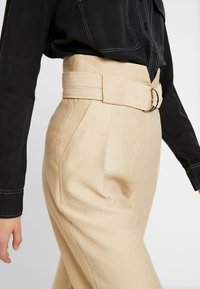 Béžové kalhoty s vysokým pasem a širokým látkovým pásem se stříbrnou sponou. Obsahují boční kapsy a hladkou texturu. Černý top s viditelnými knoflíky.