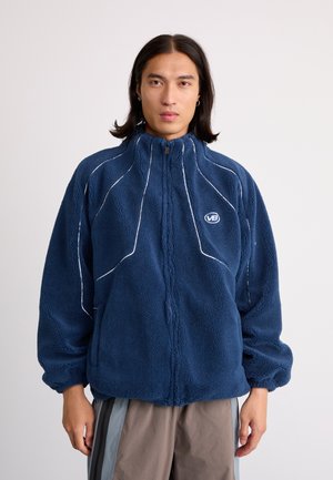 Navy blue fleece jacket with a high collar, white stitching accents, and a front zipper. Features a logo and relaxed fit with elastic cuffs.
