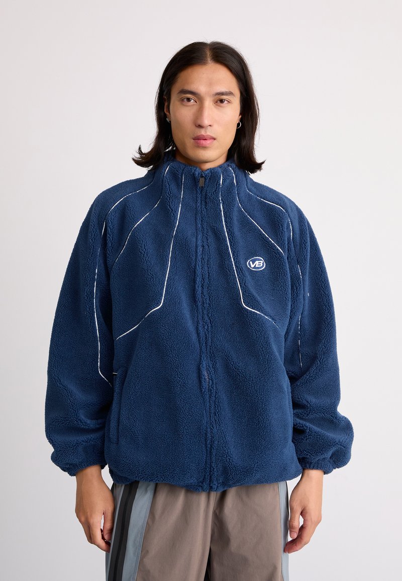 Navy blue fleece jacket with a high collar, white stitching accents, and a front zipper. Features a logo and relaxed fit with elastic cuffs.