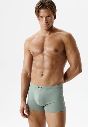 Muskularny mężczyzna o jasnej karnacji i krótkich brązowych włosach, ubrany w dopasowane bokserki w kolorze szałwii, stojący na tle jednolitej białej ściany.