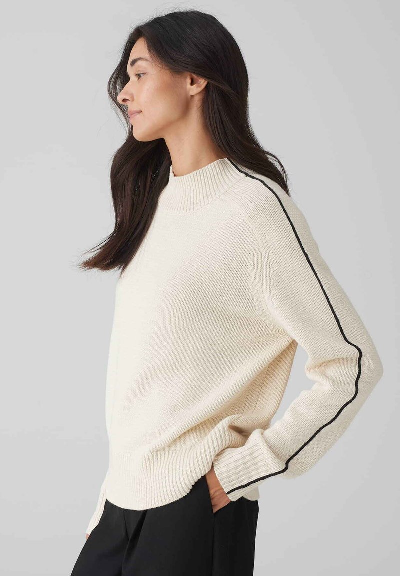 Cremfarbener Pullover mit geripptem Ausschnitt und Rippbündchen, versehen mit einem schwarzen Streifen an jedem Ärmel. Getragen mit dunklen Hosen, die eine entspannte Passform zeigen.