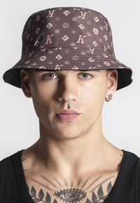 Hnedý klobúk s širokým okrajom a svetloružovým logom vzorom, vyrobený z hladkej tkaniny. Obsahuje široký okraj a prešité detaily okolo základne.