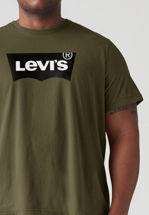 Mężczyzna w oliwkowo-zielonym t-shircie z czarno-białym logo Levi’s® na piersi, z krótkimi rękawami i widocznym tatuażem na prawej ręce.
