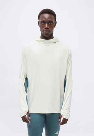 Mladý muž v svetlo béžovej mikine s dlhým rukávom a tyrkysových legínach, stojaci pred jednoduchým bielym pozadím.