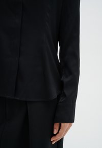 Svart satinblus med en slät yta, knappade manschetter och en lös nederkant som delvis visar en svart kjol. Ren, minimalistisk design.