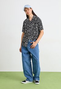 Camicia a maniche corte a righe bianche e nere, pantaloni in denim blu rilassati, sneakers nere con dettagli bianchi e un cappellino azzurro chiaro.