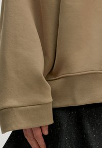 Beige sweatshirt met ribgebreide manchetten, die een soepele textuur en een relaxte pasvorm toont. De achtergrond heeft een donkere stof met spikkels.