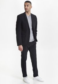Chaqueta y pantalones de traje negros combinados con una camiseta gris claro de cuello en V. Zapatillas blancas completan el conjunto. Tejido suave, diseño ajustado.